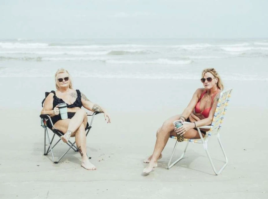 Two adult women in bikinis relax on beach chairs with drinks on a sandy beach near the ocean under an overcast sky.