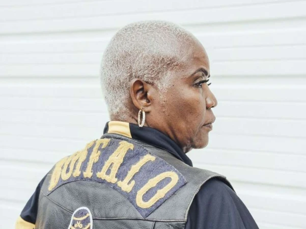 Profile portrait of an elderly Black woman with short gray hair wearing a Buffalo patch leather vest, looking off to the side against a white background.