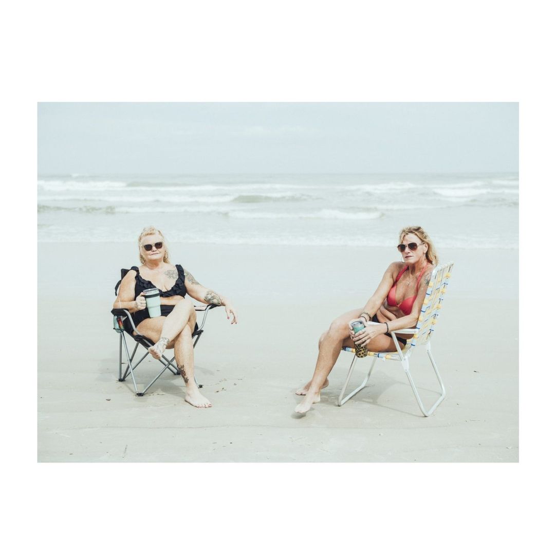 Two adult women wearing sunglasses and bikinis sit in beach chairs at the ocean shore on an overcast day in an eye-level shot.