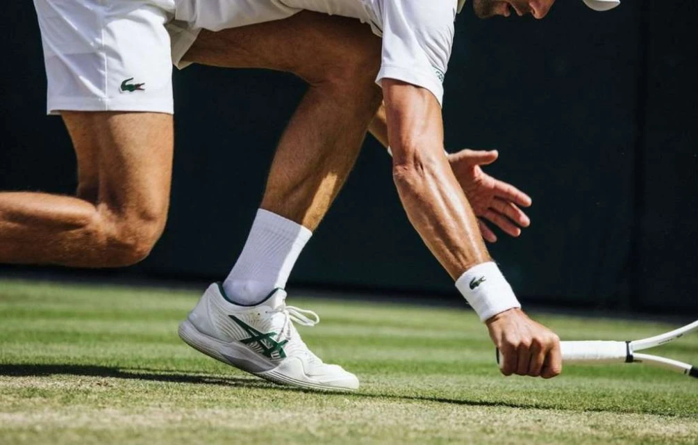 Close-up of a tennis player on a grass court, crouched low with one hand on the court and the other holding a tennis racket.