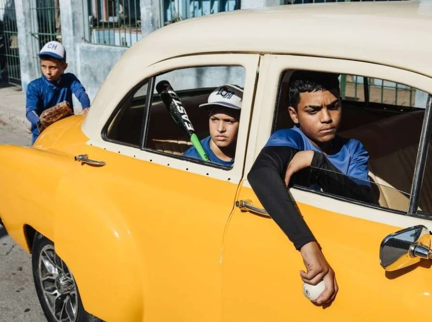 Three young baseball players in uniform pose near a yellow classic car on a city street, creating a nostalgic and sporty scene.