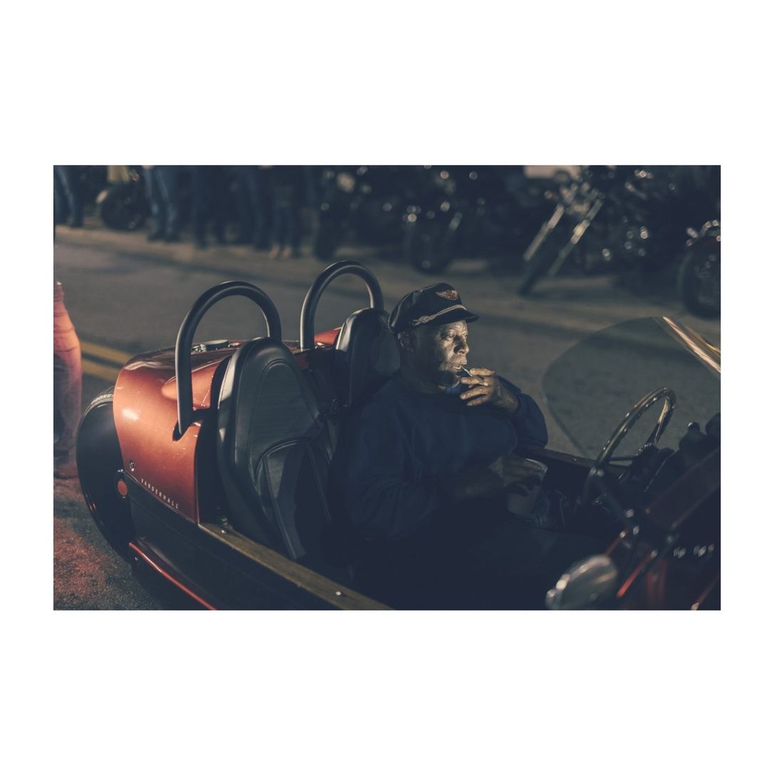 Adult male in open-top red car, vintage design, leather interior, dark background with motorcycles. Driver with cap, possible cigar.