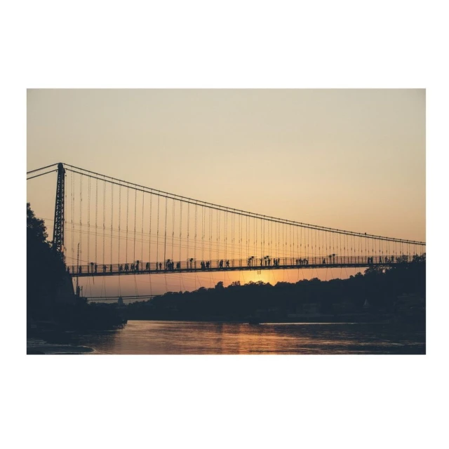 Suspension bridge over a body of water at sunset, with silhouetted figures walking across the bridge, creating a scenic landscape.