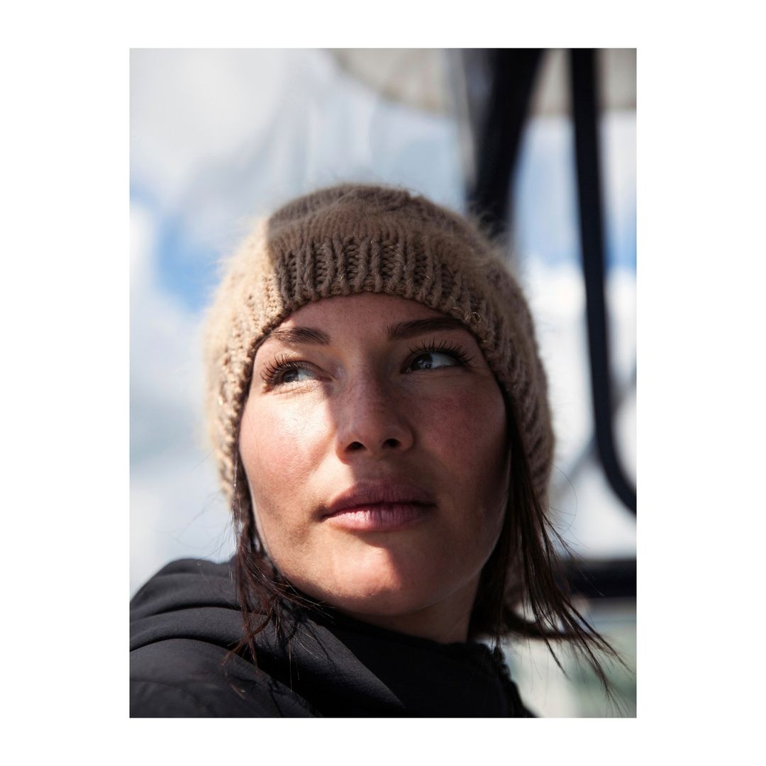 Close-up shot of a brunette woman wearing a knitted hat, looking up and away, in an outdoor setting with sky and clouds visible.