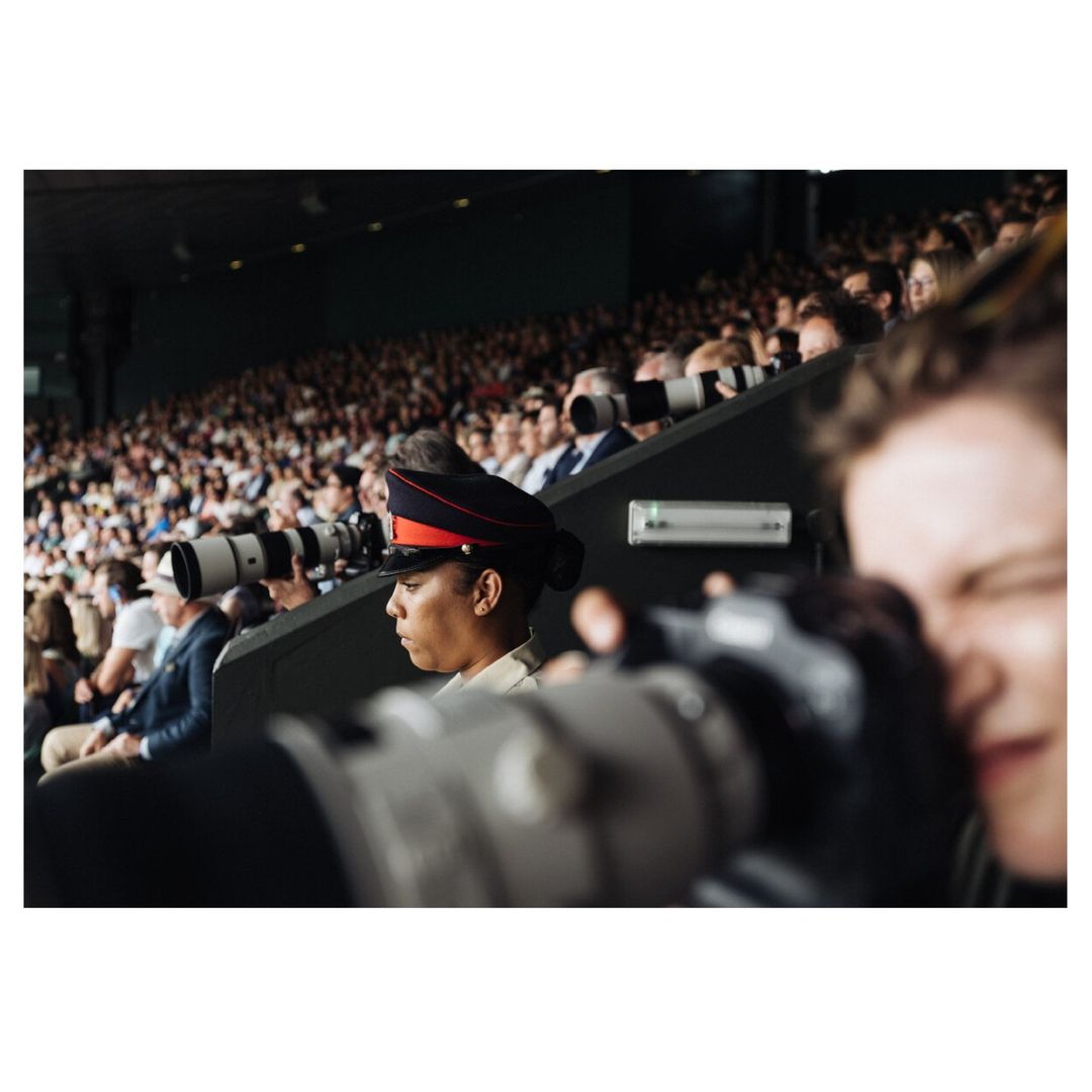 Crowd scene at a stadium with a military guard in dress uniform and photographers with long lenses capturing the event.