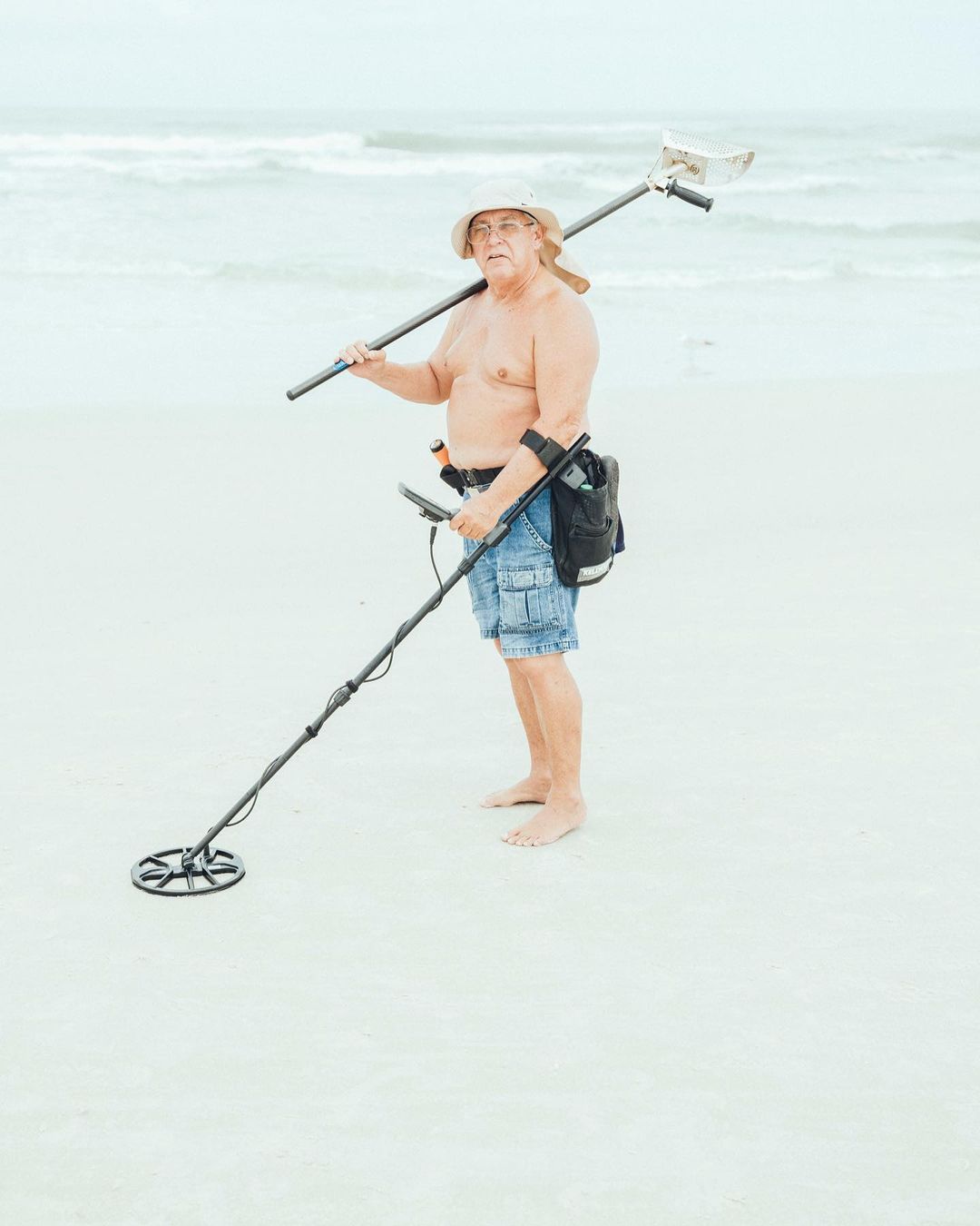 Elderly man wearing a hat metal detecting on a sandy beach with ocean waves in the background on a bright, overcast day.