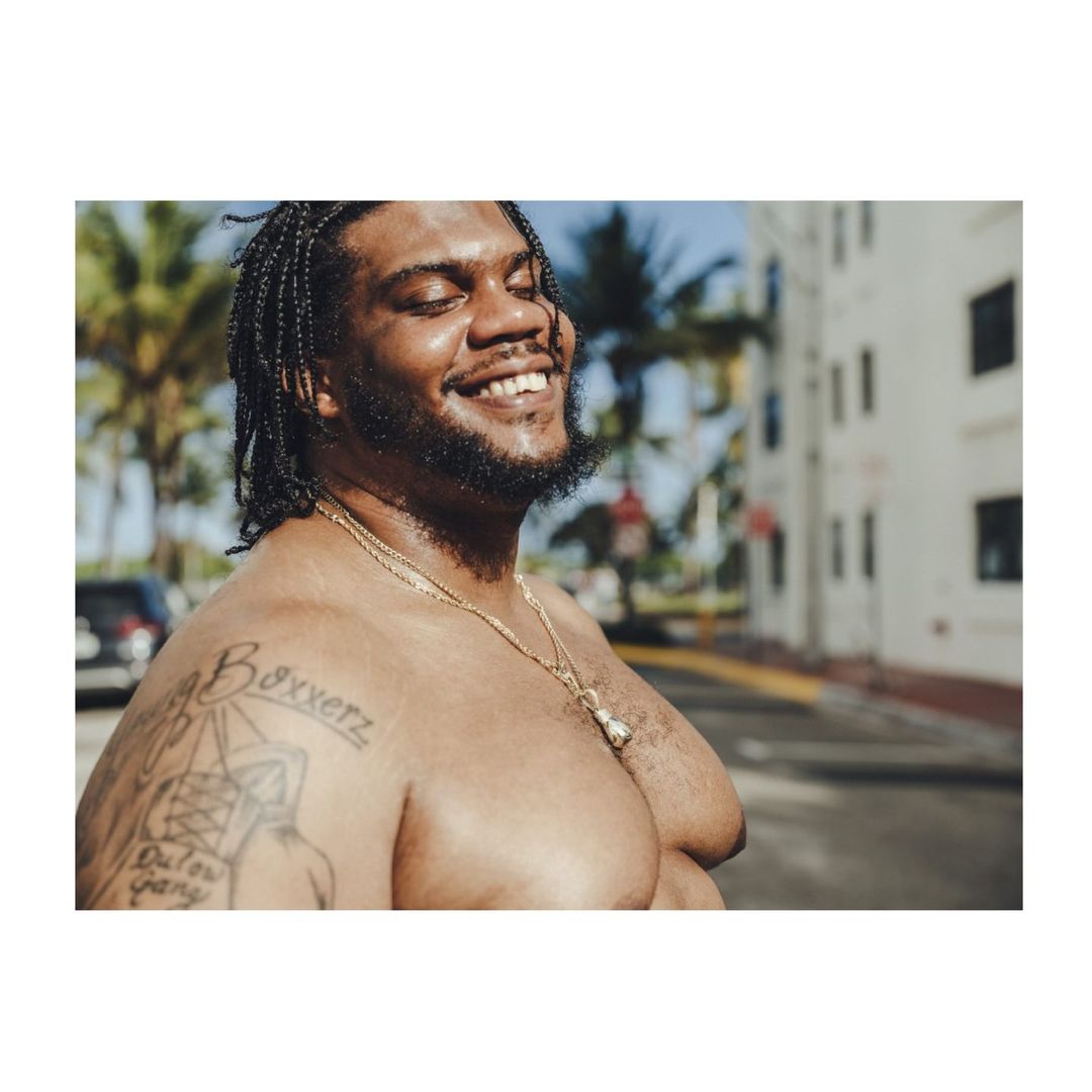 Smiling Black man with braids stands shirtless outdoors in front of white buildings and palm trees, wearing a gold chain necklace.