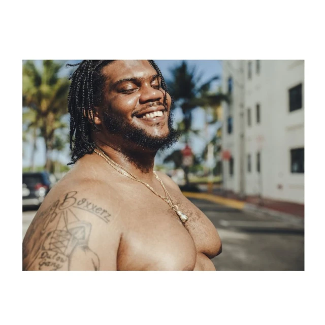 Smiling Black man with braids stands shirtless outdoors in front of white buildings and palm trees, wearing a gold chain necklace.