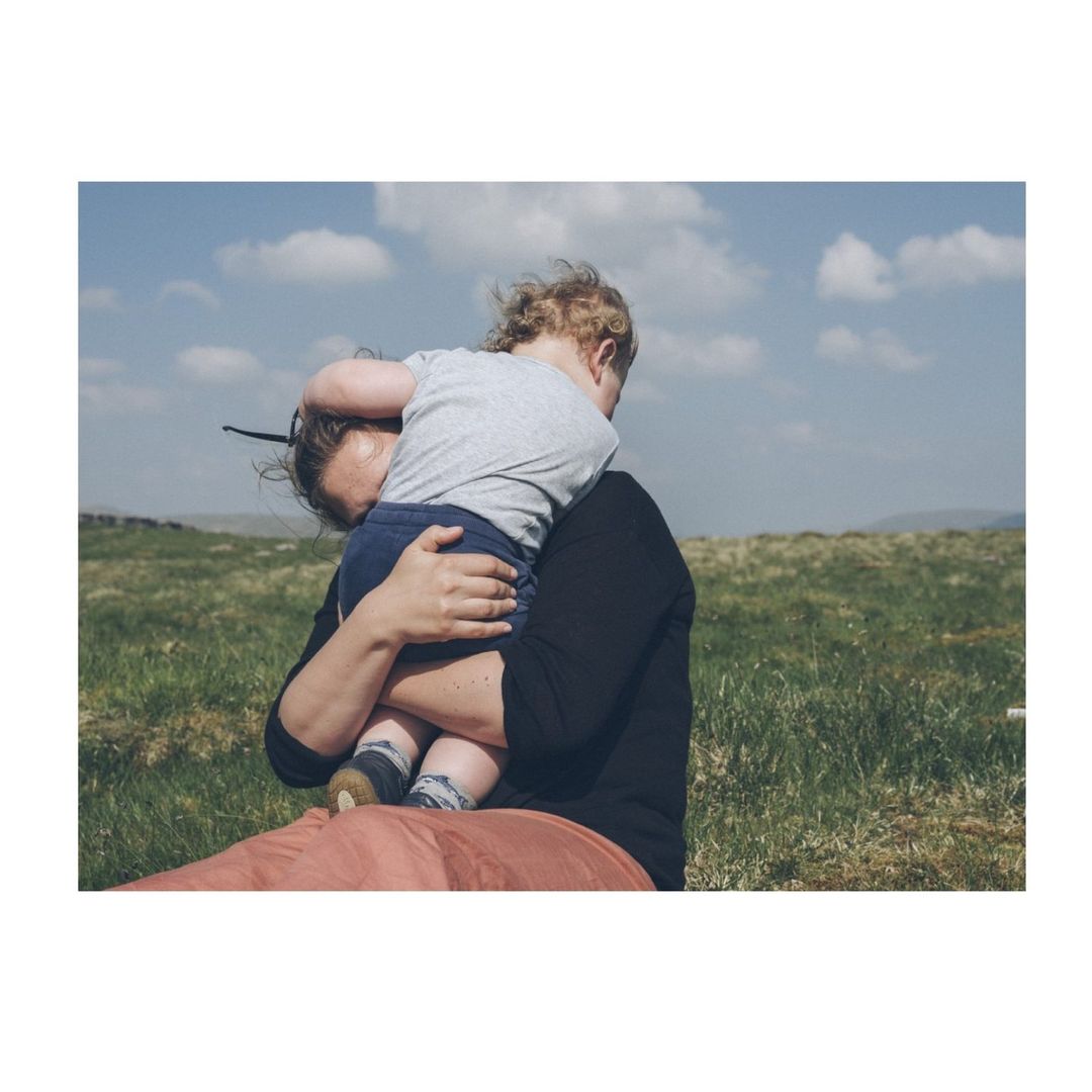 An adult holding a child in an outdoor grassy field under a blue sky with scattered white clouds.