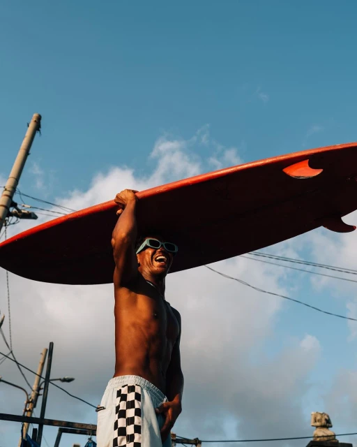Smiling African man carries surfboard above head, wearing sunglasses, set against a vibrant blue sky and urban backdrop.