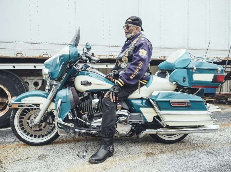 Older adult male seated on a parked turquoise and white Harley-Davidson motorcycle next to a tractor-trailer truck.