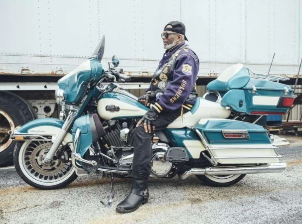 Older adult male seated on a parked turquoise and white Harley-Davidson motorcycle next to a tractor-trailer truck.