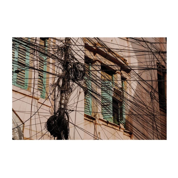Dense web of overhead power lines and tangled cables crisscrossing in front of a weathered building with green shutters