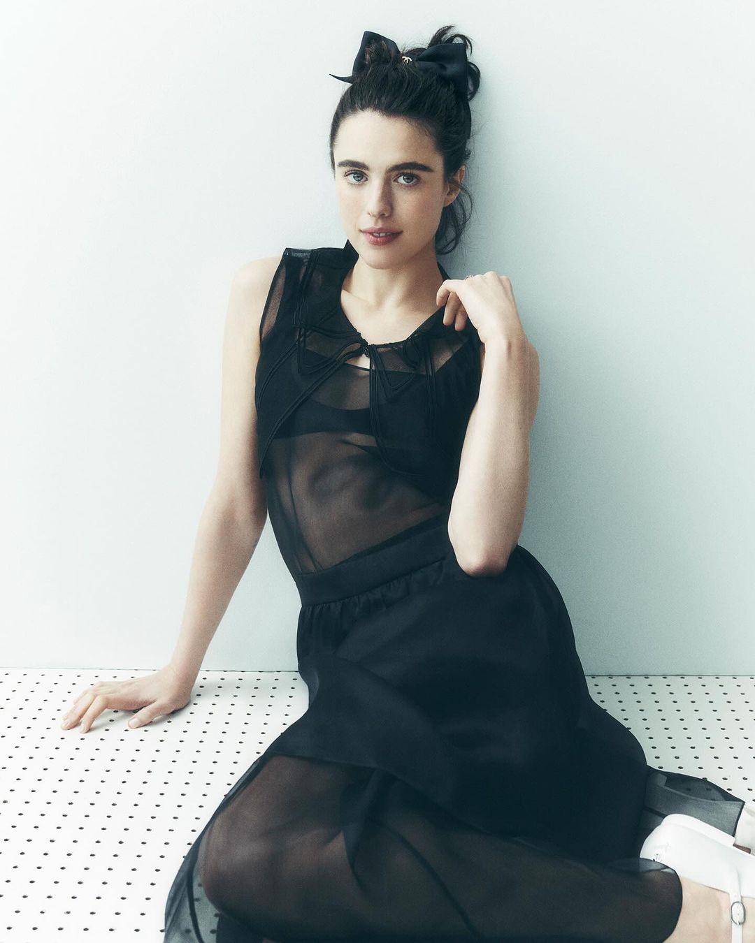Portrait of a woman with dark hair in a sheer black dress sitting on a polka dot surface in front of a white wall, looking at the camera.