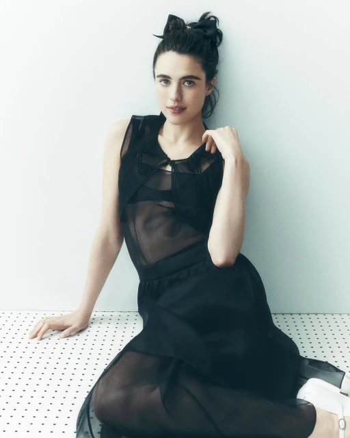 Portrait of a woman with dark hair in a sheer black dress sitting on a polka dot surface in front of a white wall, looking at the camera.