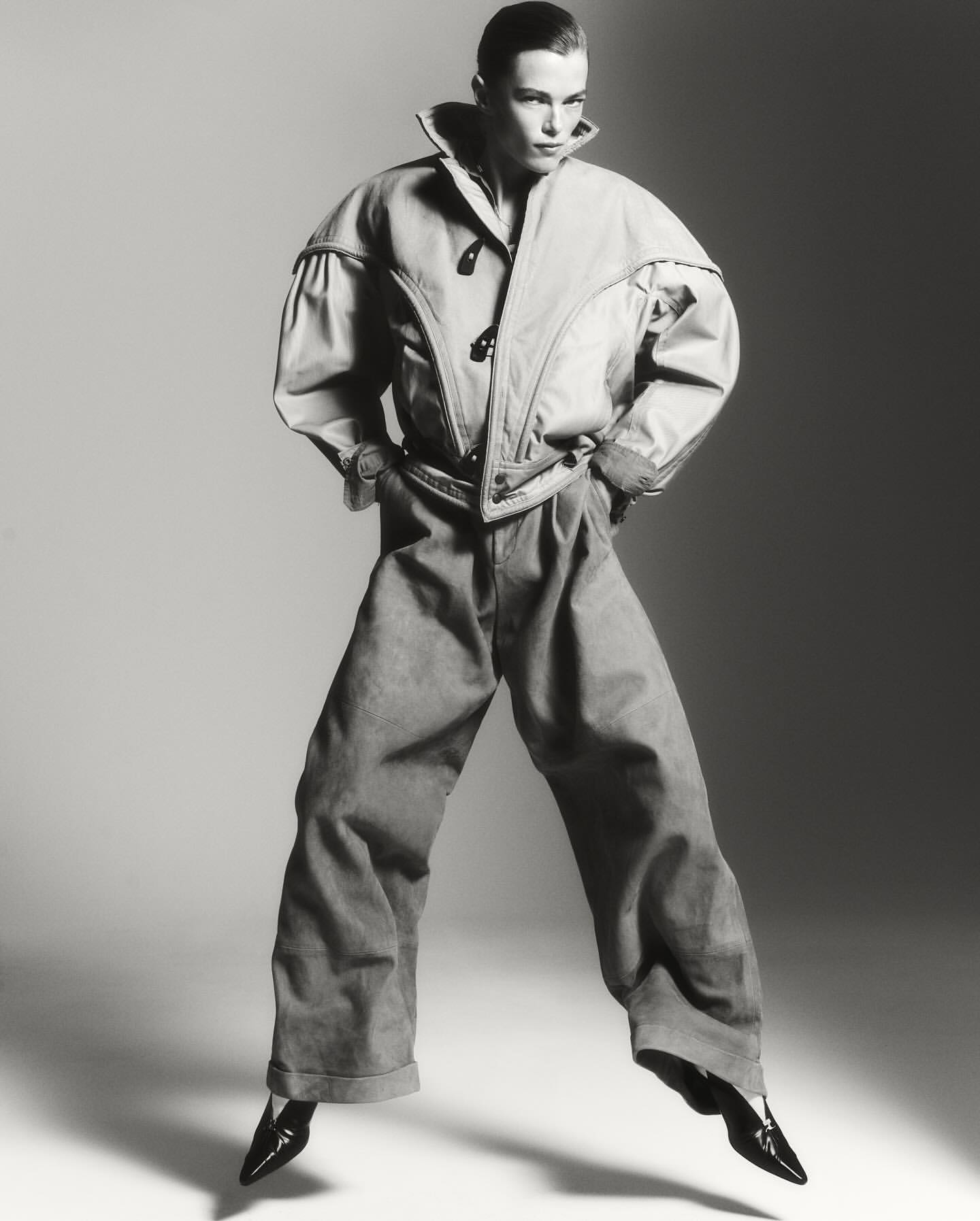 Black and white fashion shot of an adult woman with hands in pockets, wearing oversized jacket and pants, with a light grey background.