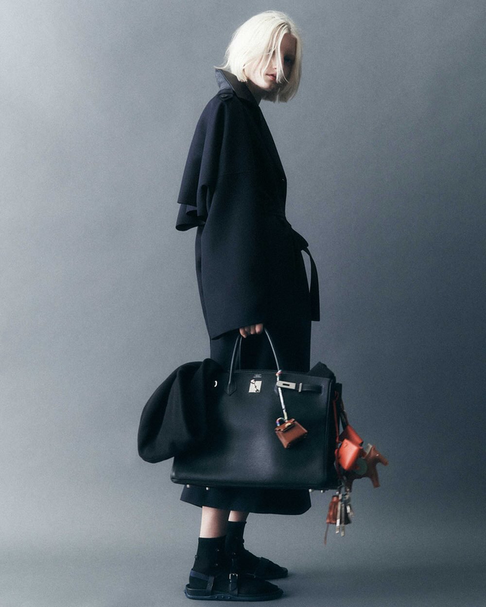 Stylish woman in a dark coat holding a black leather Birkin bag with dangling charms, posing in a studio with a grey backdrop.