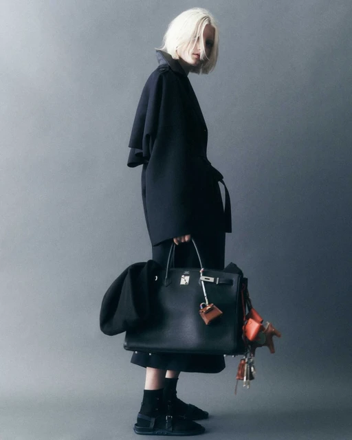 Stylish woman in a dark coat holding a black leather Birkin bag with dangling charms, posing in a studio with a grey backdrop.