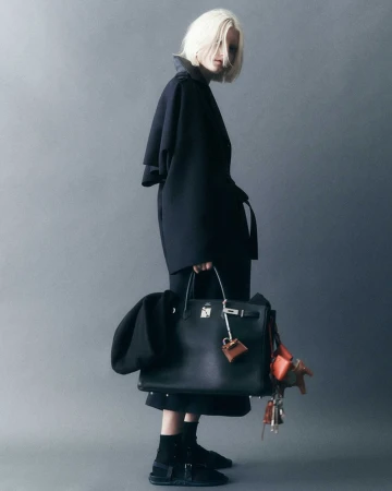 Stylish woman in a dark coat holding a black leather Birkin bag with dangling charms, posing in a studio with a grey backdrop.