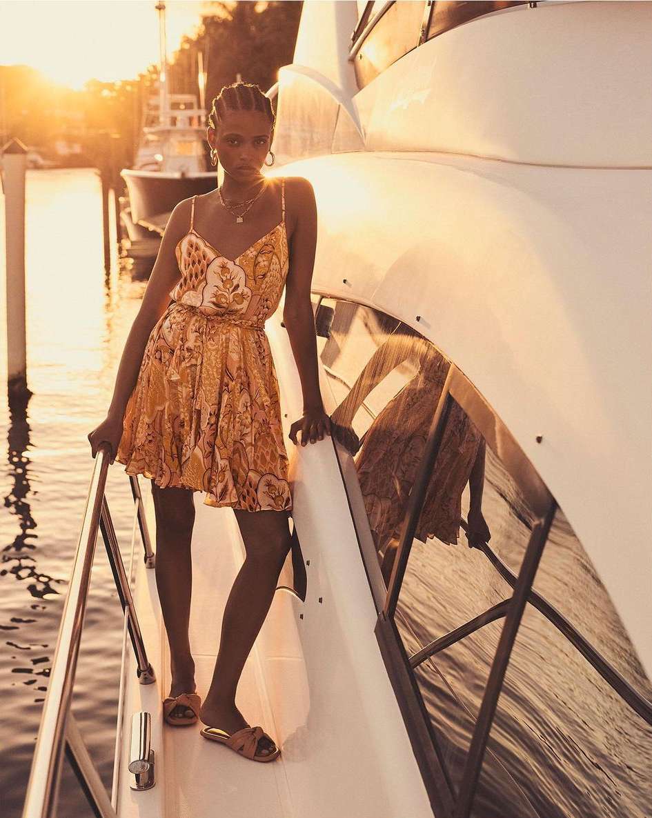 Elegant woman in a patterned dress and sandals posing on a white yacht at sunset, exuding high-end fashion and luxury travel vibes.
