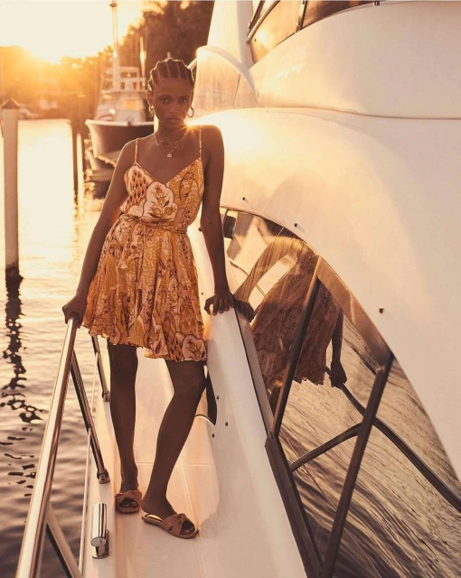 Elegant woman in a patterned dress and sandals posing on a white yacht at sunset, exuding high-end fashion and luxury travel vibes.