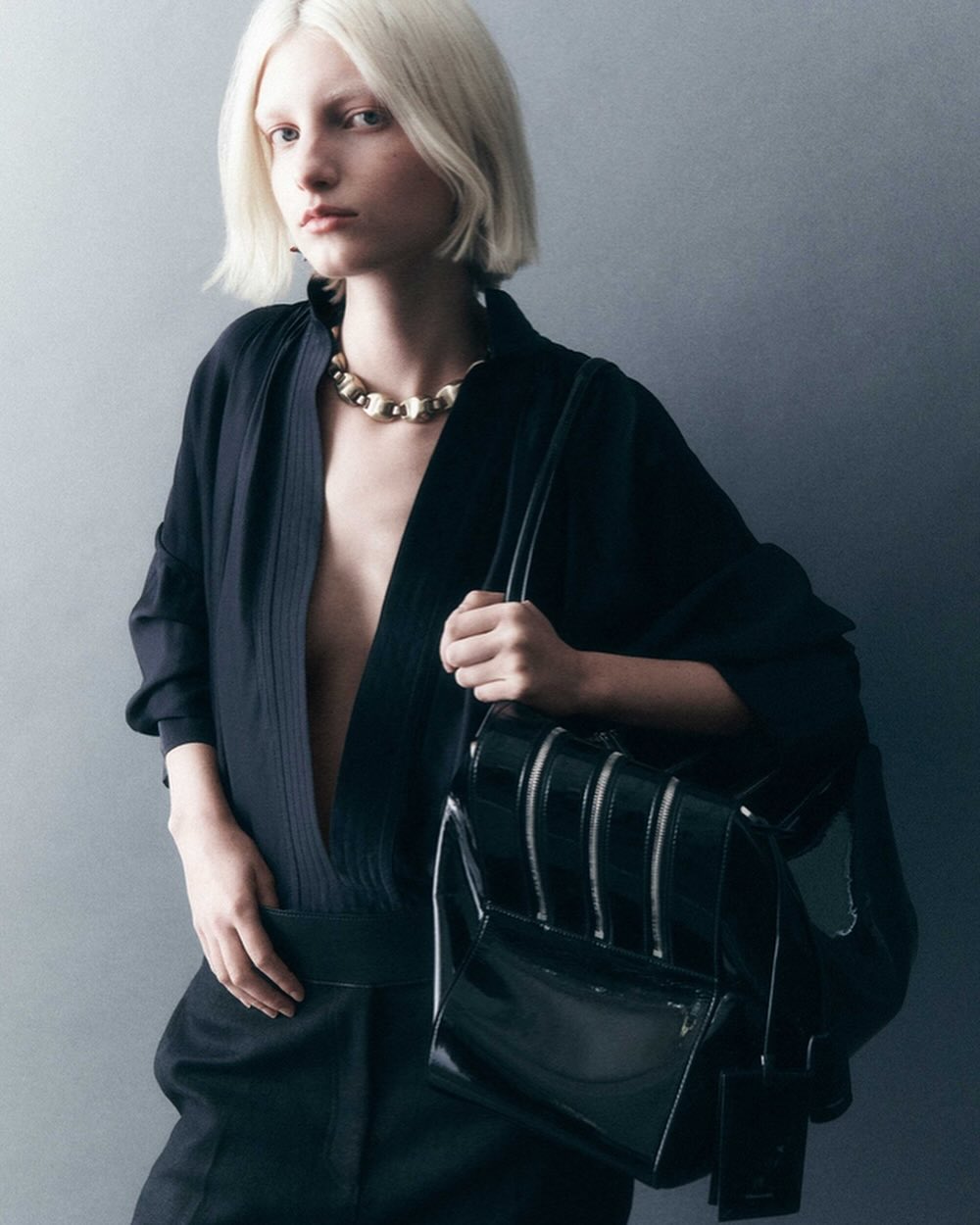 Portrait of a blonde woman wearing a black top and gold necklace holding a glossy black handbag against a grey wall