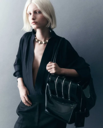 Portrait of a blonde woman wearing a black top and gold necklace holding a glossy black handbag against a grey wall