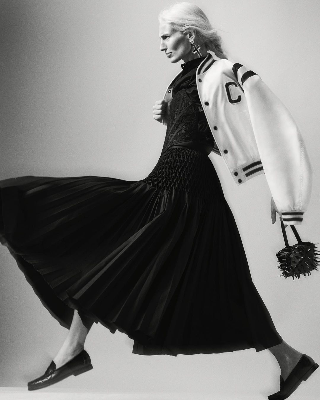 Monochrome portrait of a woman in a black dress and white jacket, striding against a light background, holding a spiky handbag.