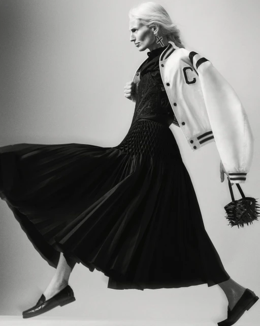 Monochrome portrait of a woman in a black dress and white jacket, striding against a light background, holding a spiky handbag.