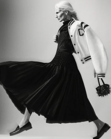 Monochrome portrait of a woman in a black dress and white jacket, striding against a light background, holding a spiky handbag.