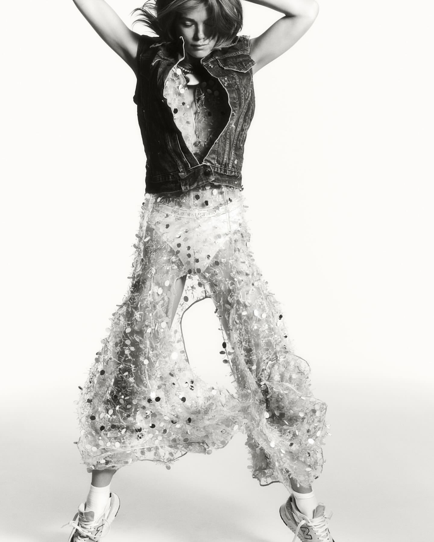 A woman jumps in a studio shot wearing a denim vest, patterned top, and sheer pants adorned with circular embellishments, adding movement.