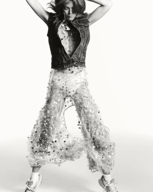 A woman jumps in a studio shot wearing a denim vest, patterned top, and sheer pants adorned with circular embellishments, adding movement.