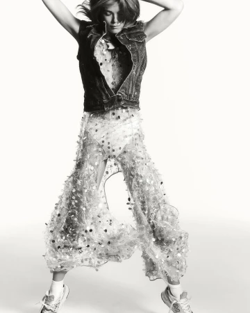 A woman jumps in a studio shot wearing a denim vest, patterned top, and sheer pants adorned with circular embellishments, adding movement.