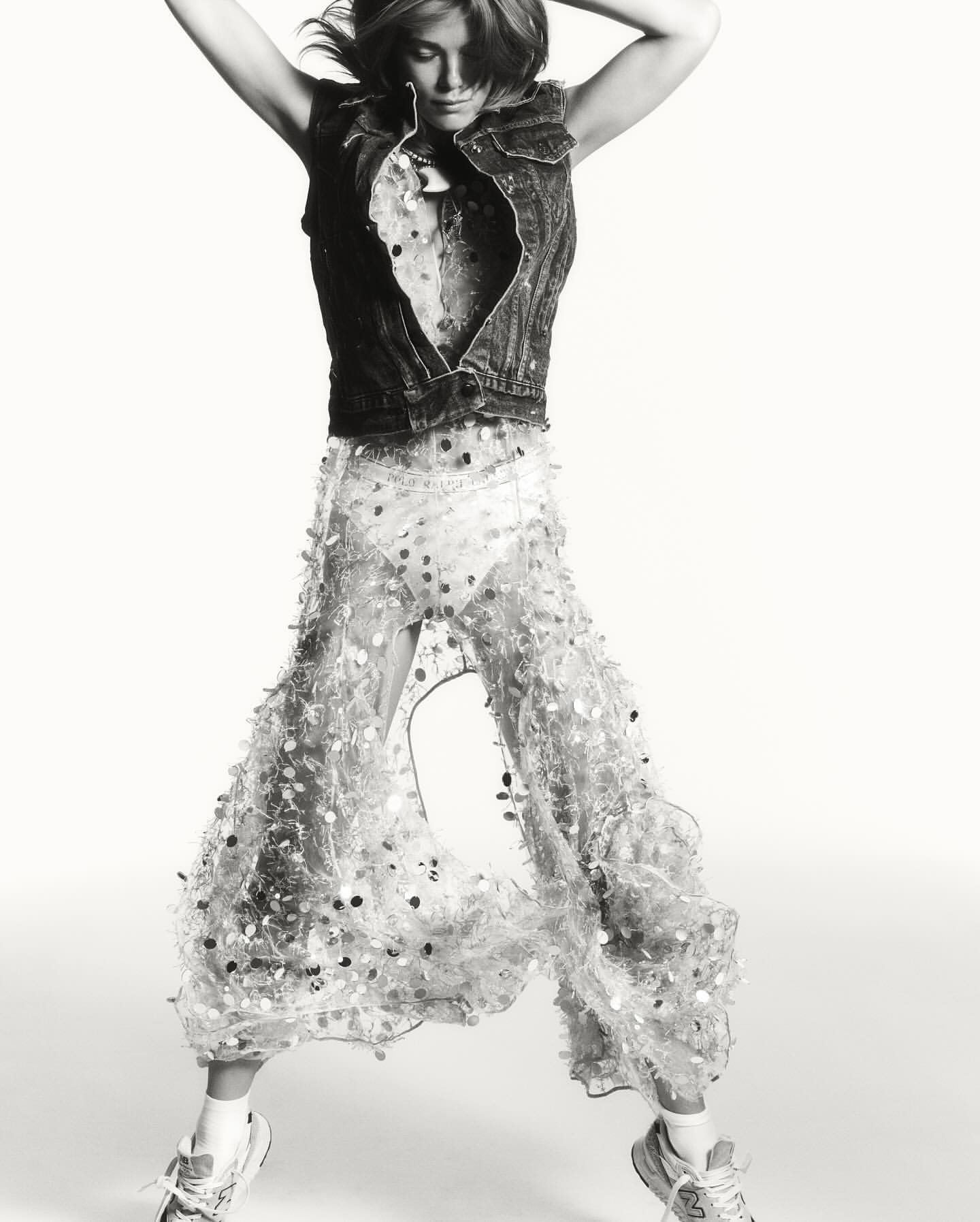 Monochrome fashion shot of a woman in mid-air wearing a denim vest and sequined pants against a stark white backdrop in a studio setting.