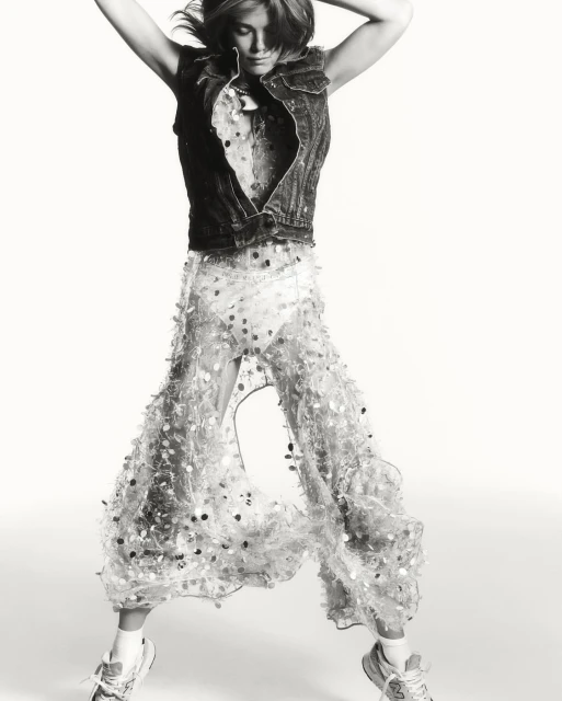 Monochrome fashion shot of a woman in mid-air wearing a denim vest and sequined pants against a stark white backdrop in a studio setting.