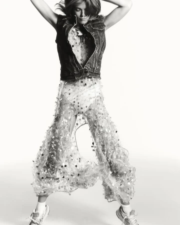 Monochrome fashion shot of a woman in mid-air wearing a denim vest and sequined pants against a stark white backdrop in a studio setting.
