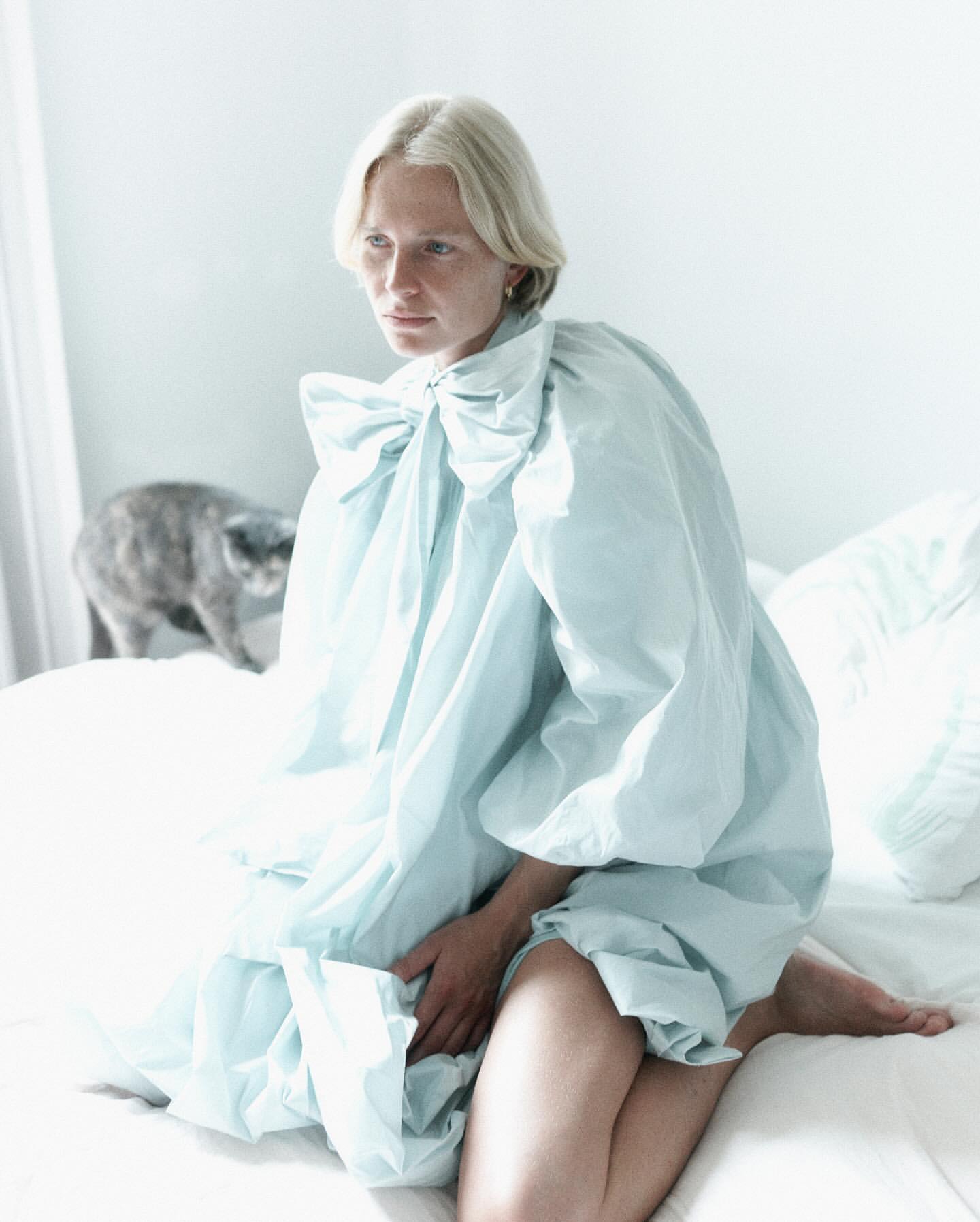 Blonde woman wearing a pale blue ruffled blouse kneeling on a white bed with a grey cat in the background.