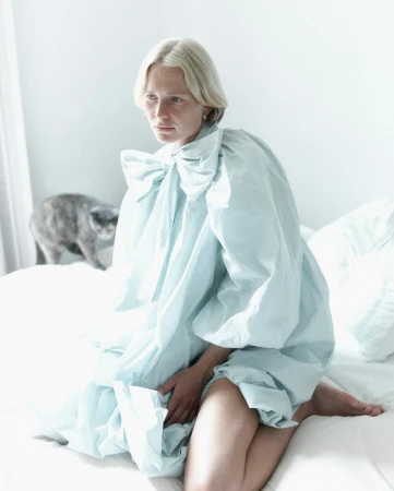 Blonde woman wearing a pale blue ruffled blouse kneeling on a white bed with a grey cat in the background.