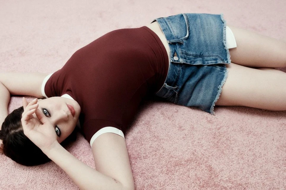 Young woman with dark hair lying on a pink carpet wearing a maroon t-shirt and denim mini skirt, gazing at the camera.