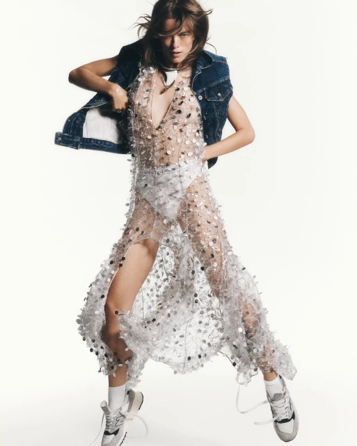 Fashionable woman in a sheer, sequined dress and denim vest posing in a dynamic stance against a bright white backdrop.
