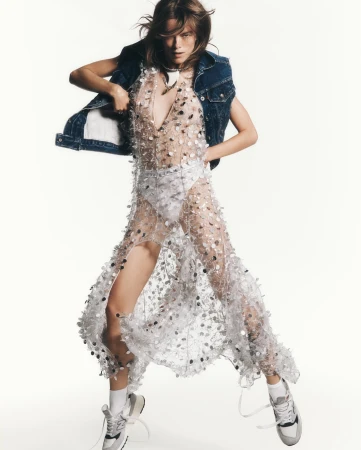 Fashionable woman in a sheer, sequined dress and denim vest posing in a dynamic stance against a bright white backdrop.