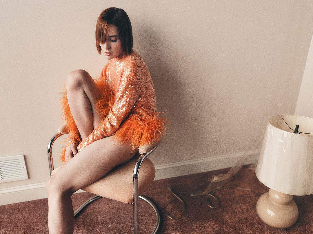 Elegant woman with short red hair poses while sitting on a chair, wearing a sequined orange feather trim top, in a minimalist indoor setting.