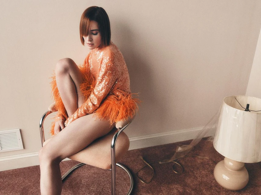 Elegant woman with short red hair poses while sitting on a chair, wearing a sequined orange feather trim top, in a minimalist indoor setting.