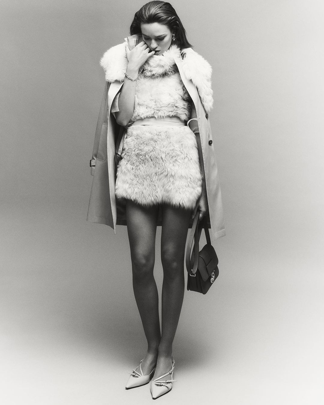 Monochrome full shot of a stylish woman in faux fur clothing, a light beige trench coat, white heels, and a small black purse in a studio.