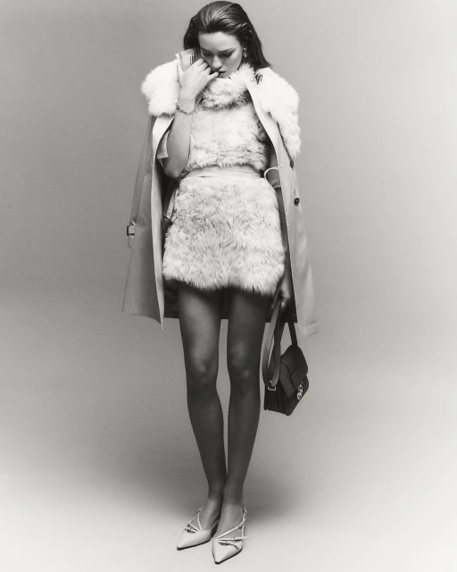 Monochrome full shot of a stylish woman in faux fur clothing, a light beige trench coat, white heels, and a small black purse in a studio.