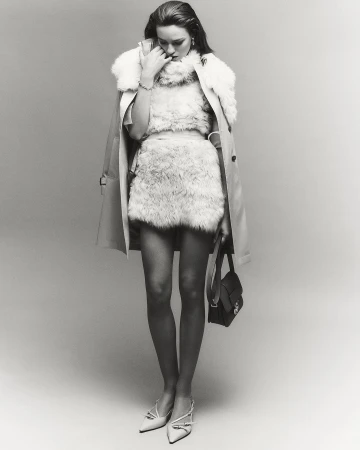 Monochrome full shot of a stylish woman in faux fur clothing, a light beige trench coat, white heels, and a small black purse in a studio.