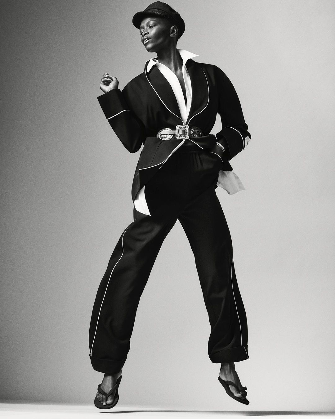 Elegant Black woman in chic black pantsuit, white shirt, and cap, is striking a pose in mid-air in a fashion photography studio.