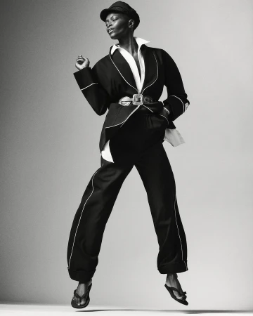 Elegant Black woman in chic black pantsuit, white shirt, and cap, is striking a pose in mid-air in a fashion photography studio.