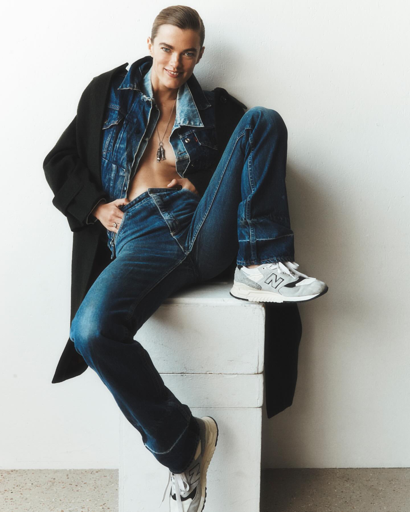 Fashionable adult male in a denim jacket and jeans, posing while sitting on a white box, wearing New Balance sneakers and a black coat.