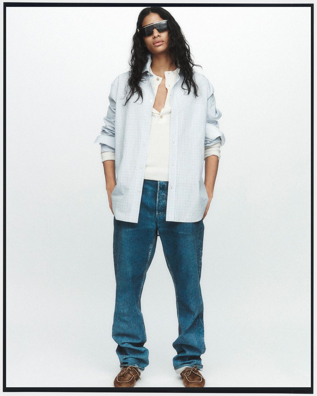 Man in unbuttoned blue striped shirt, white t-shirt, blue jeans, and brown shoes stands in a full shot against a white backdrop.